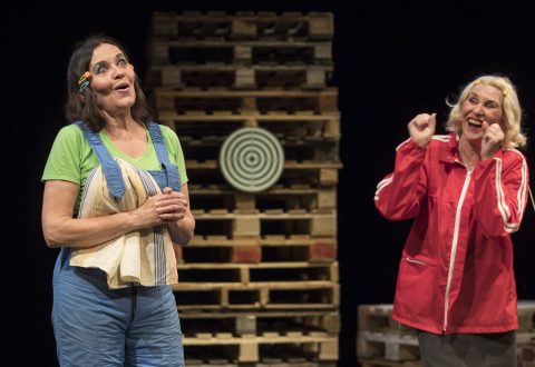 Kaksi naista esiintyy lavalla pinottujen puisten kuormalavojen edessä; toisella on sininen haalari ja hän hymyilee kädet ristissä, kun taas toinen punatakkinen nainen elehtii kiihkeästi nyrkeillä kasvojensa vieressä.