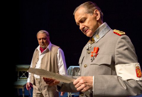 Lavalla esiintyy kaksi vanhempaa miestä, joista toinen on pukeutunut sotilasunivormuun ja lukee paperista, kun taas toinen, liiviin ja huiviin pukeutunut mies katsoo huolestuneena. Tausta on tumma ja lavasteet ovat vähäiset.