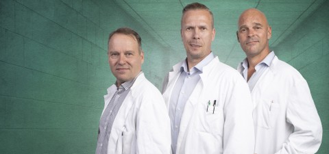 Kolme valkoisiin laboratoriotakkeihin pukeutunutta miestä seisoo vierekkäin käytävällä, jonka seinät ovat sävyltään tiilenpunaiset, ja katsoo luottavaisesti kameraan. Kahdella on lyhyet hiukset, ja yksi on kalju; kaikki vaikuttavat ammattilaisilta tai tiedemiehiltä.