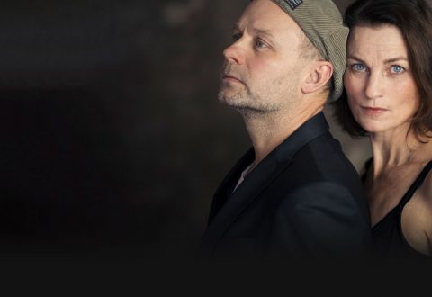 Mies, jolla on lippalakki ja tumma takki, seisoo lyhyet ruskeat hiukset omaavan naisen edessä, jolla on musta toppi. Molemmat katsovat vakavasti kameraan tummaa, sumeaa taustaa vasten.