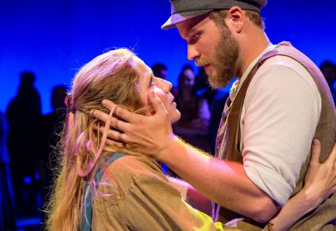 Mies pitelee hellästi naisen kasvoja, kun he katsovat toisiaan intensiivisesti lavalla, maalaispukuihin pukeutuneina, sinisellä valaistulla taustalla ja ihmisten siluetteja heidän takanaan.