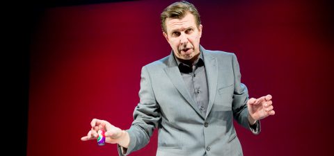 Harmaaseen pukuun ja tummaan paitaan pukeutunut mies elehtii molemmilla käsillään puhuessaan lavalla, joka on kuvattu punaisella taustalla.