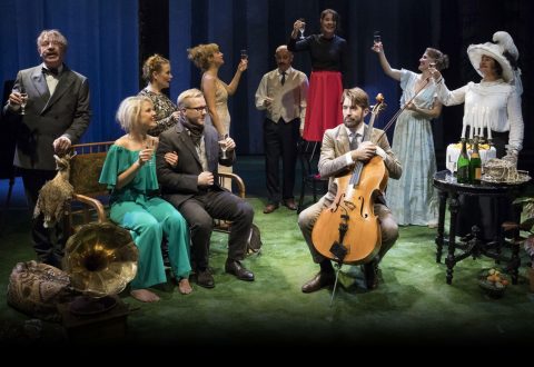 Joukko viralliseen ja vintage-tyyliseen pukeutumiseen pukeutuneita ihmisiä kohottaa maljoja juhlallisessa maljapuheessa. Yksi mies istuu edessä ja soittaa selloa, ja muut seisovat tai istuvat hänen ympärillään ruoholla ja klassisella rekvisiitalla koristellulla lavalla.