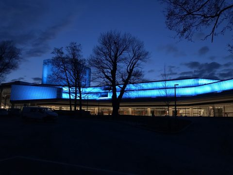 Moderni rakennus, joka on valaistu sinisillä valoilla iltahämärässä, etualalla näkyy paljaita puita ja yläpuolella on pimeä taivas. Valkoinen pakettiauto on pysäköity sisäänkäynnin lähelle.