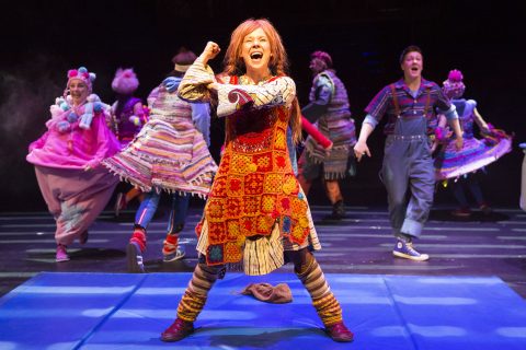 Värikkääseen, tilkkutäkkimäiseen pukuun pukeutunut esiintyjä laulaa ja elehtii energisesti lavalla, ja taustalla on eloisa joukko tanssijoita kirkkaissa, omituisissa asuissa.