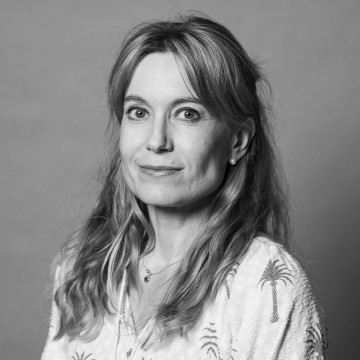 Mustavalkoinen muotokuva naisesta, jolla on pitkät, aaltoilevat hiukset ja vaalea pusero, jossa on palmukuvioita. Hän on kameraan päin, hymyilee hieman ja seisoo tavallista taustaa vasten.