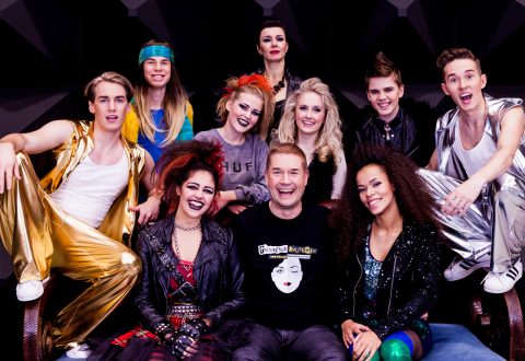 Kymmenen värikkäisiin ja näyttäviin asuihin pukeutuneen nuoren aikuisen ryhmä poseeraa yhdessä ja hymyilee innostuneesti. Useimmilla on rohkea kampaus ja kirkas meikki, mikä luo energisen ja teatraalisen tunnelman.