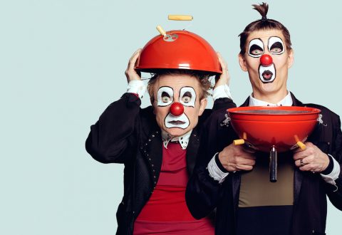 Kaksi valkoisella kasvomaalilla ja punaisella nenällä varustettua klovnia poseeraa punaisen grilliritilän kanssa; toisella on kansi hattuna, kun taas toinen pitää pohjaa rintansa edessä. Molemmilla on yllättyneet ilmeet.