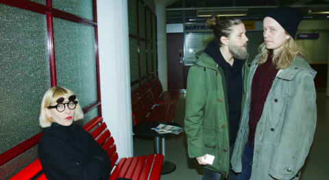 Pyöreälasipäinen nainen istuu punaisella penkillä kädet ristissä ja katsoo kameraan, kun taas kaksi vihreään takkiin pukeutunutta miestä seisoo lähekkäin keskustelemassa odotustilan taustalla.