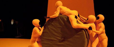 Viisi keltaisiin kokovartalopukuihin ja mustiin hanskoihin pukeutunutta ihmistä kiipeilee ja kerääntyy suuren, pyöreän, vihreällä pehmustetun esineen ympärille keltaisella lavalla, jonka taustalla näkyy valonheitin.