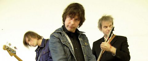 Kolme kitaraa soittavaa miestä seisoo vaaleaa taustaa vasten. Keskellä olevalla miehellä on farkkutakki ja hän katsoo suoraan kameraan, kun taas kaksi muuta, hieman epäselvästi erottuvat, keskittyvät soittimiinsa.