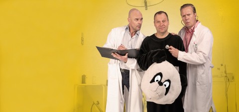 Kolme miestä seisoo keltaisen taustan edessä; kahdella on valkoiset laboratoriotakit, ja heillä on leikepöytä ja stetoskooppi kädessään, ja he tutkivat kolmatta miestä, joka on pukeutunut mustaan karhupukuun ja pitää pandan päähinettä käsissään.