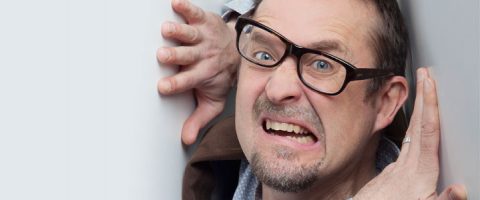 Silmälasipäinen mies näyttää ahdistuneelta ja painautuneelta valkoista seinää vasten, irvistellen silmät suurina ja kulmakarvat koholla, samalla kun hänen kätensä painuvat pintaa vasten.