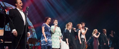 Ryhmä muodollisesti pukeutuneita esiintyjiä seisoo rivissä lavalla ja laulaa mikrofoneihin värikkäiden lavavalojen alla.