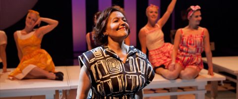 Rohkeaan mustavalkokuvioiseen mekkoon pukeutunut nainen hymyilee itsevarmasti keskellä näyttämöä, kun taas kolme värikkäisiin vintage-asuihin pukeutunutta naista istuu ja poseeraa valkoisilla penkeillä hänen takanaan näyttämövalojen alla.