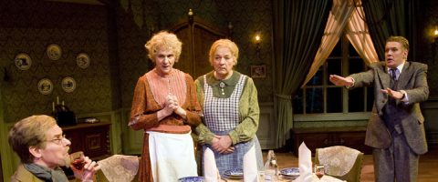 Neljä näyttelijää esittää dramaattisen kohtauksen vanhassa ruokasalissa; kaksi naista seisoo huolestuneina pöydän ääressä, kun taas yksi mies elehtii kiireesti ja toinen juo lasista. Tunnelma on jännittynyt.