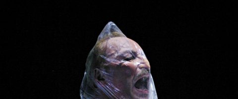 Viiksekäs henkilö näyttää huutavan tai haukkovan henkeään, kun hänen päänsä on kääritty tiukasti kirkkaaseen muoviin mustaa taustaa vasten.