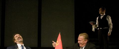 Kaksi pukumiestä istuu pöydän ääressä naureskellen, ja heidän välissään on pieni Neuvostoliiton lippu. Taustalla seisoo mustaan liiviin ja rusettisolmioon pukeutunut tarjoilija, jolla on tarjotin kädessään ja joka tarkkailee kohtausta. Tausta on tumma.