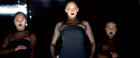 Kolme mustiin asuihin ja dramaattiseen meikkiin pukeutunutta esiintyjää, joilla on suuret silmät ja intensiiviset kasvonilmeet, seisovat tummaa taustaa vasten luoden vaikuttavan teatteritunnelman.