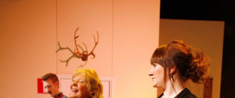 Kolme ihmistä sisätiloissa, etualalla kaksi naista laulamassa tai puhumassa ja taustalla mies, heidän takanaan näkyy seinään kiinnitetty sarvikoriste. Kohtaus on lämpimästi valaistu.