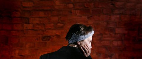 Lyhythiuksinen ja otsapantainen henkilö on tiiliseinää vasten, käsi poskella ja selkä pääosin kameraan päin. Valaistus on hämärä ja tunnelma mietiskelevä.