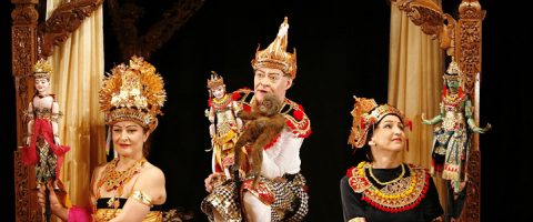 Kolme esiintyjää koristeellisissa perinteisissä balilaisissa puvuissa ja päähineissä istuu koristellulla näyttämöllä ja pitää kädessään nukkeja. Keskimmäisellä esiintyjällä on myös apina. He näyttävät olevan keskellä teatteriesitystä.