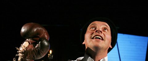 Mustaan pipoon ja valkoiseen paitaan pukeutunut mies pitelee ruskeaa nyrkkeilyhanskaa ja katsoo ylöspäin iloisin ilmein tummaa taustaa vasten.