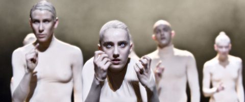 Viisi esiintyjää, joilla on vaalea meikki ja tiukat puvut, seisovat dramaattisen valaistuksen alla, ottavat voimakkaita, ilmeikkäitä asentoja ja tuijottavat eteenpäin luoden surrealistisen ja teatraalisen ilmapiirin.