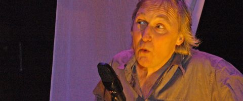 Vaaleatukkainen mies, jolla on yllättynyt ilme, puhuu mikrofoniin lavalla, jota valaisee violetti ja keltainen valaistus.