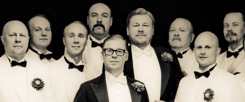 Kahdeksan miestä, jotka ovat pukeutuneet virallisiin valkoisiin smokkeihin, joissa on mustat rusetit ja kukkakimput, seisovat yhdessä tumman taustan edessä ja poseeraavat ryhmäkuvaa varten.