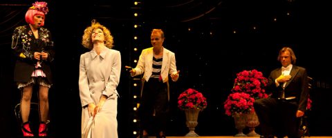 Neljä näyttelijää esiintyy näyttämöllä dramaattisin ilmein, kirkkaiden näyttämövalojen ja suurten vaaleanpunaisten kukkamaljakoiden ympäröimänä. Yhdellä on valkoinen puku, toisella smokki, ja yhdellä on kirkkaan vaaleanpunainen tukka ja rohkea asu.