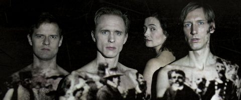 Neljä vakavan näköistä ihmistä, kolme miestä ja yksi nainen, seisovat lähekkäin tummaa taustaa vasten, ja heidän paljaalle ylävartalolleen ja kasvoilleen heijastetaan mustavalkoisia historiallisia kuvia.