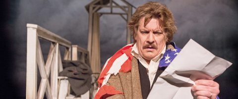 Viiksekäs mies lukee intensiivisesti isoa paperia Amerikan lippuun sonnustautuneena ja seisoo puisen rakennelman edessä, jonka taustalla on pilvinen taivas.