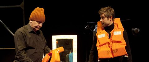 Kaksi miestä seisoo lavalla; toisella on ruskea neulehattu ja hänellä on kädessään oranssi pelastusliivi, kun taas toinen, jolla on musta paita ja oranssi pelastusliivi, katsoo häntä. Taustalla näkyy valaistu peili.
