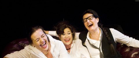 Kolme valkoisiin paitoihin pukeutunutta naista istuu lähekkäin tummanpunaisella sohvalla ja nauraa iloisesti. Tausta on tumma, ja kaikki kolme vaikuttavat iloisilta ja rennoilta, joista yhdellä on silmälasit ja löysästi sidottu solmio.