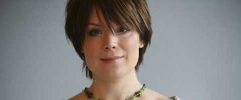 Henkilö, jolla on lyhyet ruskeat hiukset ja otsatukka, hymyilee pehmeästi, yllään vihreä helmikaulakoru ja vaalea toppi, harmaalla taustalla.