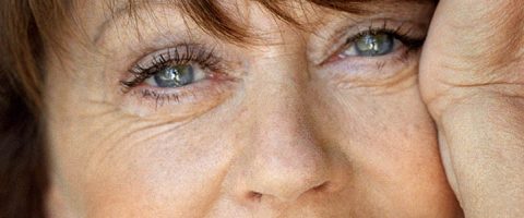 Lähikuva vanhemman naisen kasvoista, joissa näkyy hänen siniset silmänsä, kevyet rypyt ja osa hänen kädestään lepää hänen poskeaan vasten.