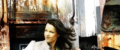 Pitkä tummahiuksinen nainen, jolla on vaalea toppi, seisoo ruostuneiden, sääntelemien puuovien edessä ja katsoo sivulle, kun tuuli puhaltaa hänen hiuksiaan.