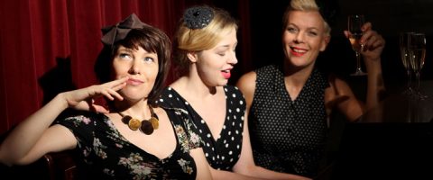 Kolme vintage-tyyliseen mekkoon pukeutunutta naista istuu yhdessä punaisen verhon edessä. Yksi nainen hymyilee ja pitelee juomaa kädessään, toinen laulaa, ja kolmas näyttää mietteliäältä ja koskettaa huuliaan.