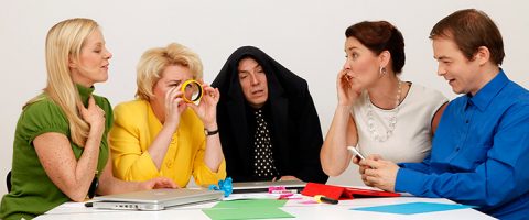 Viisi aikuista istuu pöydän ääressä paperien, kannettavien tietokoneiden ja toimistotarvikkeiden kanssa. He näyttävät vitsailevan - yksi nainen tutkii teippirullaa, toisella on takki päänsä päällä, ja ryhmä on sitoutunut ja nauraa.