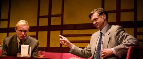 Kaksi pukumiestä istuu hämärässä huoneessa, jossa on punaiset istuimet ja geometrinen puuseinä. Toinen mies näyttää vakavalta, kun taas toinen osoittaa sormellaan ja elehtii kuin osoittaisi jotain heidän keskustelunsa aikana.