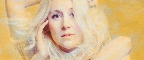 Pitkät, vaaleat hiukset omaava nainen poseeraa kädet kasvojensa lähellä keltaista taustaa vasten ja katsoo suoraan kameraan pehmeällä, neutraalilla ilmeellä.