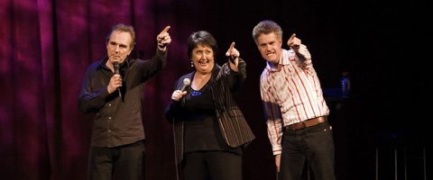 Lavalla seisoo kolme ihmistä, joilla on mikrofonit kädessään ja jotka hymyilevät ja osoittavat kohti yleisöä. Taustalla on tumma violetinpunainen verho.