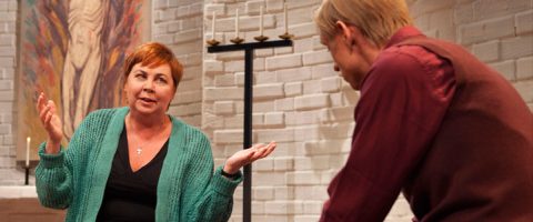 Vihreään neuletakkiin pukeutunut nainen elehtii käsillään puhuessaan punaruskeaan paitaan ja liiviin pukeutuneelle miehelle. He ovat sisätiloissa, joissa on valkoiset tiiliseinät, kynttilöitä ja maalaus taustalla.