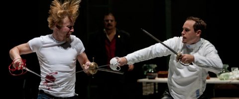 Kaksi miestä käy lavalla kiihkeää miekkailua; toisella on valkoinen miekkailutakki ja toisella verinen valkoinen t-paita. Kolmas henkilö tarkkailee taustalla.