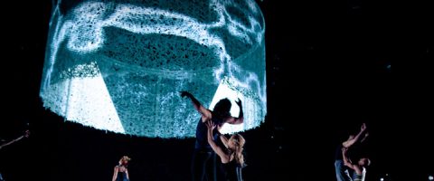 Esiintyjät tanssivat pimeällä näyttämöllä suuren, valaistun, pyöreän rakenteen alla, johon on heijastettu abstrakteja valokuvioita. Tunnelma on dramaattinen ja taiteellinen.