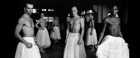 Kuusi paidatonta miestä pukeutuu valkoisiin balettitutuihin ja seisoo itsevarmasti hämärässä huoneessa katsoen kohti kameraa. Tunnelma on dramaattinen ja taiteellinen, voimakkaat varjot ja pehmeä valo.