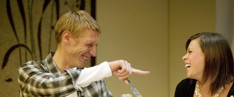 Ruudulliseen paitaan pukeutunut mies osoittaa leikkisästi hymyilevää ruskeatukkaista naista, ja molemmat nauravat ja nauttivat yhdessä sisätiloissa hilpeästä hetkestä.