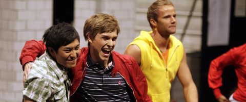 Kolme nuorta miestä hymyilee ja poseeraa yhdessä; kaksi heistä on lähellä toisiaan, toisella on punainen takki ja toisella vihreä ruutupaita, kun taas kolmas mies keltaisessa hihattomassa hupparissa seisoo taustalla.