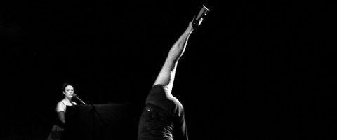 Nainen laulaa mikrofoniin vasemmalla, kun taas toinen esiintyjä, joka on enimmäkseen kuvan ulkopuolella, nostaa oikealla tummalla taustalla yhden jalan korkealle ilmaan.